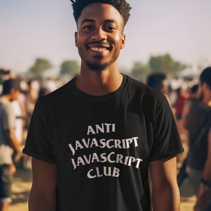 May include: A man wearing a black t-shirt with white text that reads "ANTI JAVASCRIPT JAVASCRIPT CLUB". He is smiling and standing in front of a crowd of people.