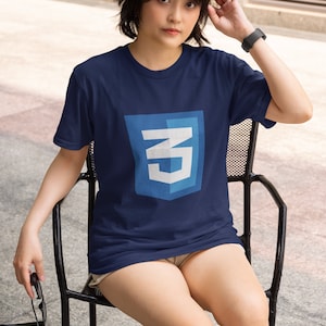May include: Navy blue t-shirt featuring a shield design with the letters "CN" in white and light blue. The person is sitting on a black chair. The shirt is a short-sleeved tee.