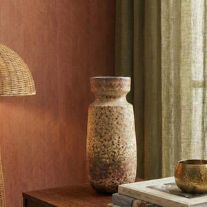 Può includere: Un vaso alto in ceramica strutturata con finitura beige e marrone macchiettata. Il vaso ha una base ampia e un collo più stretto. È appoggiato su una superficie di legno con libri e una piccola ciotola dorata. Lo sfondo presenta un muro marrone-rossastro e tende verdi.