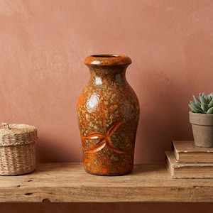 Può includere: Un vaso in ceramica marrone e arancione con superficie strutturata e un motivo a stella intagliato. Il vaso è posto su una mensola di legno con un cestino intrecciato, una pianta grassa e dei libri.