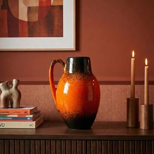 Op de afbeelding: Een grote, oranje en zwarte keramische kan met een handvat staat op een donkere houten ondergrond. Twee brandende kaarsen in houten houders en een stapel boeken staan ook op het oppervlak. Een abstract schilderij hangt op de achtergrond.