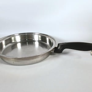 Könnte beinhalten: Eine Edelstahl-Bratpfanne mit schwarzem Griff und silbernem Aufhängerring. Die Pfanne hat eine glänzende, reflektierende Oberfläche und eine runde Form, geeignet zum Kochen.