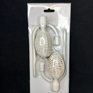 May include: White woven-style curtain tiebacks in a clear plastic package. The tiebacks have an oval shape with a woven texture and are connected by a curved, white metal piece. The package has the word "HOME" printed on it.