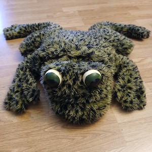 May include: A plush frog-shaped rug with a textured, mottled green and black pattern. The frog has large, round eyes with white and green details. The rug is lying on a wooden floor.