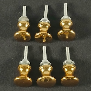 Peut inclure: Six boutons de meuble dorés avec une base arrondie et un corps en forme de sablier. Chaque bouton a une tige filetée argentée pour le montage. Les boutons sont disposés sur une surface gris foncé.