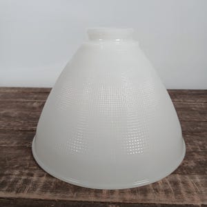 May include: A white, cone-shaped lampshade with a textured, grid-like pattern. The lampshade has a rounded base and a narrower top, designed to fit a light fixture. The shade is made of opaque glass or a similar material.