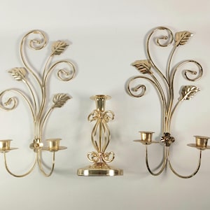 May include: A set of three gold-colored metal candle holders consisting of two wall-mounted sconces and one freestanding pillar candle stand. Each sconce features two candle cups and decorative metal scrollwork with leaf-shaped accents.