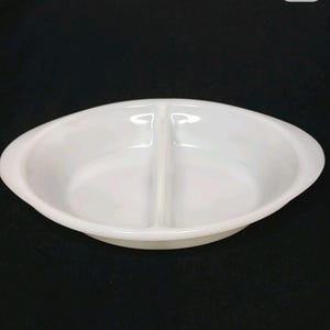 Könnte beinhalten: Eine weiße, ovale Servierschale mit Unterteilung. Die Schale besteht aus glänzendem Material und verfügt über eine mittige Trennwand, die zwei separate Fächer schafft. Sie hat abgerundete Kanten und Griffe auf beiden Seiten.