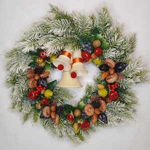 May include: A festive Christmas wreath featuring faux frosted pine branches, adorned with ceramic bells, red berries, and various dried fruits. The wreath has a circular shape and is set against a white background.