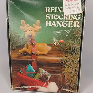 Op de afbeelding: Een kerstkoushanger met een rendierfiguur, een rode riem en een rode kous vol cadeaus. Op de doos staat "REIN STOCKING HANGER". De achtergrond bevat dennentakken.