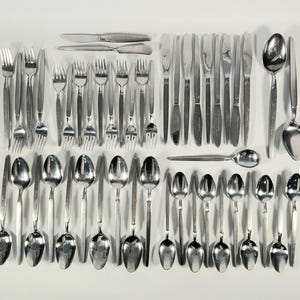 May include: A collection of stainless steel silverware, including forks, knives, and spoons, arranged on a white surface. The set includes various sizes of forks, knives, and spoons, reflecting a complete dining set.