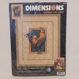 Peut inclure: Kit de point de croix compté Dimensions avec un motif de coq royal. Le kit comprend une image détaillée d'un coq aux plumes orange, bleues et marron, sur un fond vert et beige. Le kit mesure 28 x 36 cm.