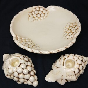 May include: Off-white ceramic serving dish and two candle holders. The dish has a scalloped edge and grape cluster designs. The candle holders are shaped like grape clusters with leaf details. The pieces have a glossy finish.