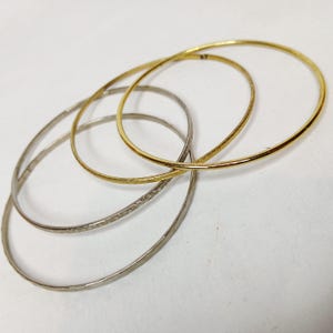 May include: Four thin bangles in gold and silver tones. Two are gold, one with a textured surface. The other two are silver, one with a textured surface. The bangles are overlapping on a white surface.