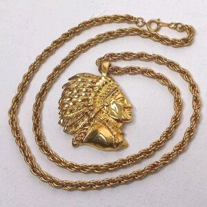 May include: A gold-toned necklace featuring a detailed pendant of a Native American chief in profile. The pendant is suspended from a thick, textured rope chain. The necklace is displayed on a white surface, highlighting the gold color.