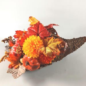 May include: A woven dark brown wicker cornucopia overflowing with artificial autumn decor. The arrangement features vibrant orange and red maple leaves, a bright yellow dahlia, small orange pumpkins, and textured golden-brown floral accents.