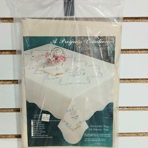 May include: A Progress Creation stamped tablecloth in a clear plastic package. The tablecloth is off-white with floral embroidery in pink, blue, and yellow. The package includes a matching napkin. The text on the package reads "Dynasty" and "Permanent Press Soil Release Finish."