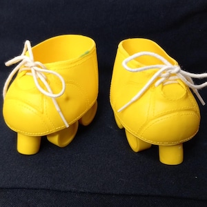 Pode incluir: Um par de sapatos de patins de plástico amarelo com cadarços brancos. Os sapatos têm um design simples e estão sobre um fundo preto.