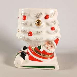 Op de afbeelding: Een keramische plantenbak in de vorm van een witte kerstboom met zilveren glitteraccenten en rode ornamenten, rustend op een basis met een kerstmanfiguur in een rood pak met zwarte laarzen, geplaatst op een witte slee met een groene rand.