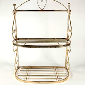 May include: A two-tier gold-toned metal wall shelf featuring a curved frame with decorative scrollwork on the sides and a heart-shaped wire accent at the top. The unit has two slatted shelves with a rounded front edge and a polished metallic finish.