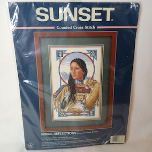 Op de afbeelding: Een Sunset kruissteekpakket. De afbeelding toont een Native American vrouw met twee poema's, omlijst door een decoratieve rand. De kit is verzegeld in een plastic verpakking. Afmeting afbeelding: 23x36cm.