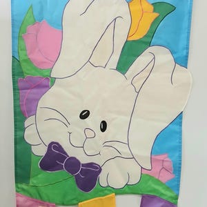 May include: Easter-themed flag with a cartoon bunny, tulips, and the text "HOPPY EASTER!". The bunny has a purple bow tie and is surrounded by pink, yellow, and purple flowers. Three small, colorful egg-shaped flags hang below.