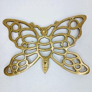 Puede incluir: Un salvamanteles de latón con forma de mariposa y diseño calado. El salvamanteles tiene un patrón detallado de círculos y líneas, y es de color dorado cálido. Las alas son simétricas y el cuerpo tiene un extremo puntiagudo. Este artículo decorativo es adecuado para la cocina.