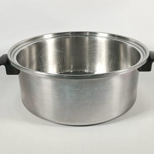Pode incluir: Uma panela de cozinha em aço inoxidável com um acabamento metálico brilhante. Possui duas alças pretas e uma abertura ampla. A panela está vazia e repousa sobre uma superfície branca. O diâmetro da panela é de cerca de 25 cm.