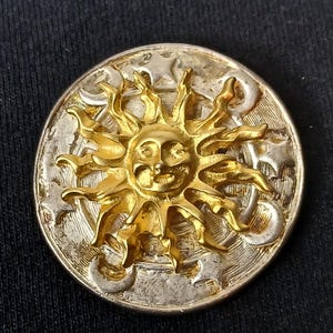 May include: A round, silver-toned button with a gold-colored sun design at the center. The sun has a smiling face and radiating rays. The button's border features stars and crescent moons, creating a celestial theme.