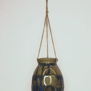 May include: A hanging ceramic planter with a brown and blue glaze. The planter has three arched openings and is suspended by a natural fiber rope. The rounded shape and color variations give it a rustic, handmade appearance.