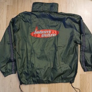 May include: An olive green windbreaker jacket featuring a high collar and long sleeves with black and white checkered side stripes. The back displays an embroidered red oval patch with the text Johnny Blaze in white and silver lettering.