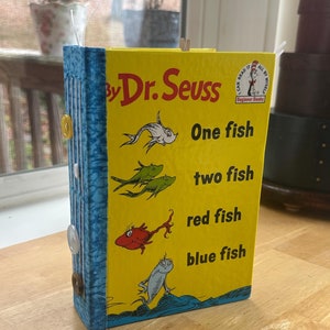 May include: A handmade book with a yellow cover featuring the title "One Fish, Two Fish, Red Fish, Blue Fish" by Dr. Seuss. The book has a blue spine with decorative buttons and a small wooden clip at the top.
