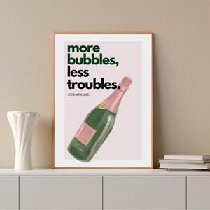 May include: A framed art print featuring a green champagne bottle with a pink label and top. The print has the words "more bubbles, less troubles" in green and black text. The word "champagne" is below the text.