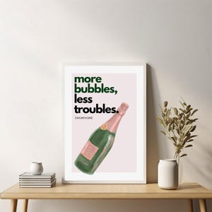 May include: A framed art print featuring a green champagne bottle with a pink label and top. The print has the text "more bubbles, less troubles." in green and black. The word "CHAMPAGNE" is below the text.