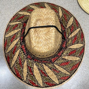 May include: A straw hat with a woven design featuring red, green and black accents. The hat has a wide brim and a black ribbon around the crown.
