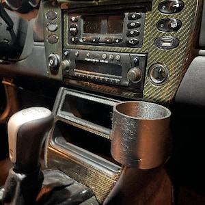 May include: A black cup holder is mounted in the center console of a car. The cup holder is made of a textured material and has a round opening. The console is made of a carbon fiber material and has a silver gear shift knob.
