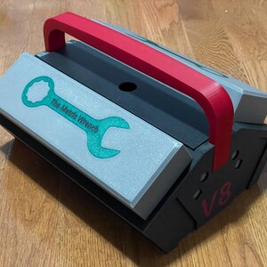 Könnte beinhalten: Eine schwarze und silberne Werkzeugbox mit einem roten Griff. Die Werkzeugbox hat ein grünes Schraubenschlüssel-Logo mit dem Text "The Metric Wrench" und die Nummer "V8" an der Seite.