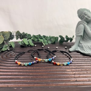May include: Three black string bracelets with colourful gemstone beads. Each bracelet has seven beads in a rainbow colour pattern. The bracelets are displayed on a wooden surface with a small green Buddha statue in the background.