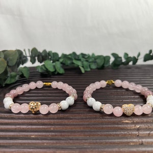 May include: Two bracelets with pink and white beads and gold heart charms. The bracelets are on a wooden surface.