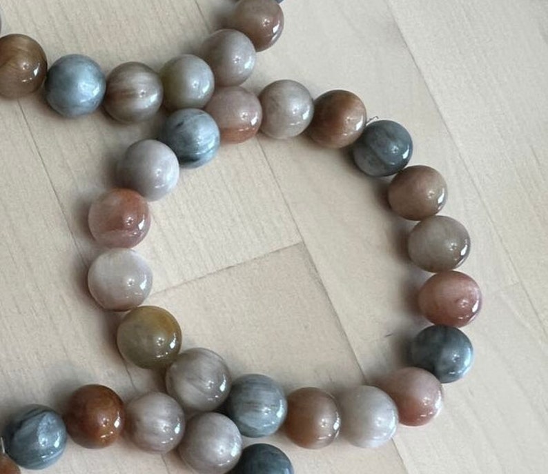 May include: A strand of sunstone beads in shades of brown, grey, and white. The beads are round and have a smooth, polished finish.
