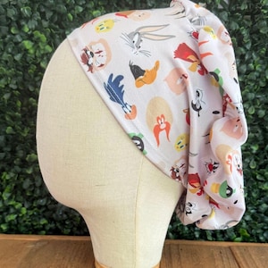 May include: A white and light pink scrub cap featuring cartoon characters like Bugs Bunny, Daffy Duck, and others. The cap is displayed on a mannequin head against a green leafy background.