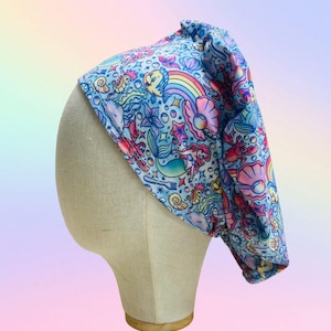 May include: A light blue scrub cap featuring a colorful mermaid-themed pattern. The design includes mermaids, seashells, rainbows, and various sea creatures in shades of pink, yellow, and blue. The cap is displayed on a mannequin head.