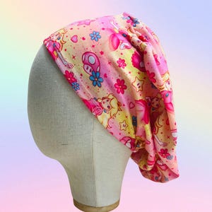 May include: A pink and yellow patterned head covering featuring cartoon characters, flowers, and stars. The fabric is wrapped around a mannequin head, showcasing the design and fit of the headwear.