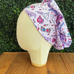 May include: A light purple and white patterned scrub cap featuring various magical symbols and icons. The cap is displayed on a mannequin head against a green background and wooden surface. The design includes glasses, lightning bolts, and hearts.