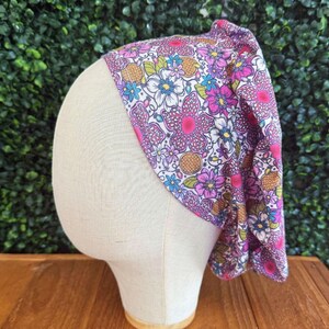 May include: A floral headband with a vibrant pattern of pink, purple, blue, and yellow flowers on a white background. The headband is displayed on a mannequin head against a green backdrop.