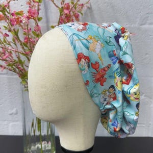 May include: A light blue scrub cap featuring a repeating pattern of characters from The Little Mermaid, including Ariel, Flounder, Sebastian, and King Triton. The fabric is gathered at the back and is displayed on a mannequin head.