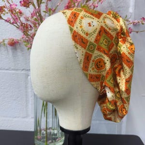 May include: A fabric scrub cap featuring a repeating diamond pattern in shades of orange, cream, and green. The print includes illustrations of jack-o'-lanterns, small ghosts, and bats, with a gathered back design for a secure and adjustable fit.