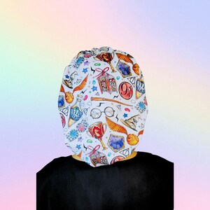 May include: White patterned cap featuring various Harry Potter-themed illustrations, including a golden snitch, potion bottles, and the Hogwarts crest. The cap is worn over a black shirt.