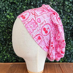 May include: A pink patterned cap with a repeating design of hearts, cherries, bows, and the word "LOVE". The cap is displayed on a mannequin head against a backdrop of green foliage and a wooden surface.
