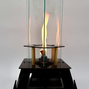 May include: A black metal and glass tabletop fireplace with a flame burning inside. The fireplace has a rectangular base and a clear glass enclosure. The text "STONE EMBER" is visible on the base.